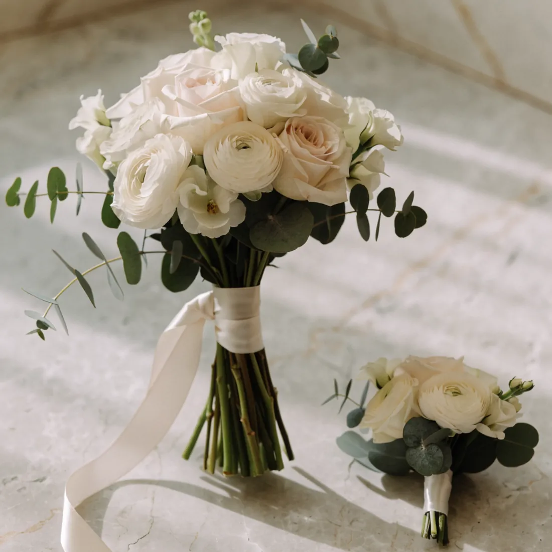 Brautstrauß aus weißen und zartrosa Rosen und Ranunkeln mit Seidenband, daneben eine Anstecknadel